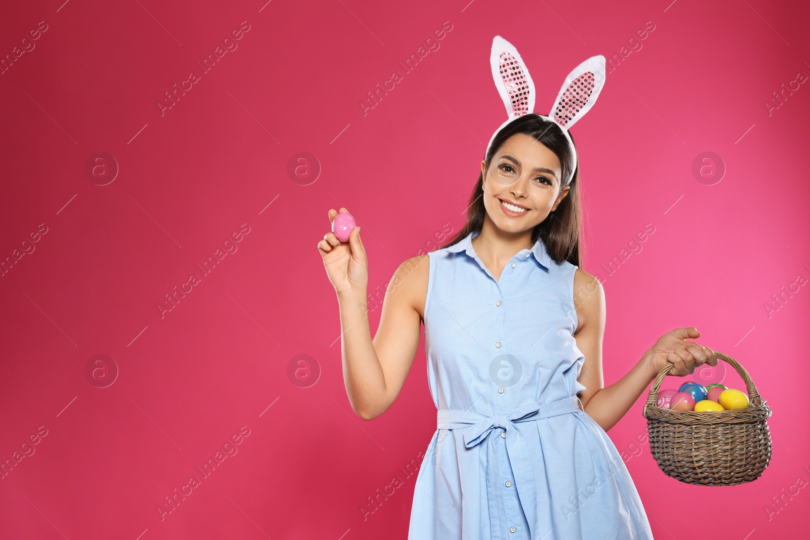 Photo of Beautiful woman in bunny ears headband holding basket with Easter eggs on color background, space for text