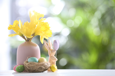 Photo of Painted Easter eggs, ceramic bunny and flowers on white table against blurred background. Space for text