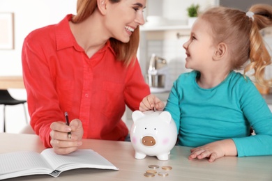 Photo of Mother and daughter putting coin into piggy bank at table indoors. Saving money