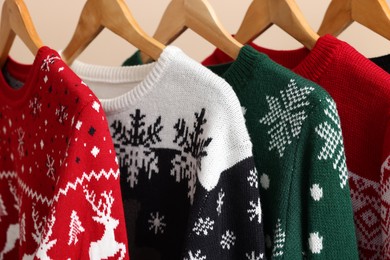 Different Christmas sweaters hanging on rack against beige background, closeup