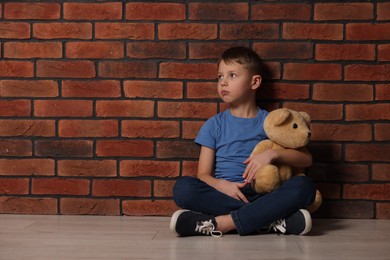 Photo of Child abuse. Boy with teddy bear sitting on floor near brick wall indoors, space for text