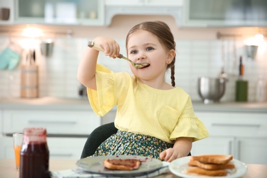 Photo of Little girl eating jam at table in kitchen