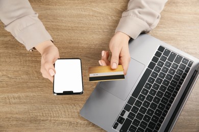 Online payment. Woman using credit card and smartphone with blank screen near laptop at wooden table, top view