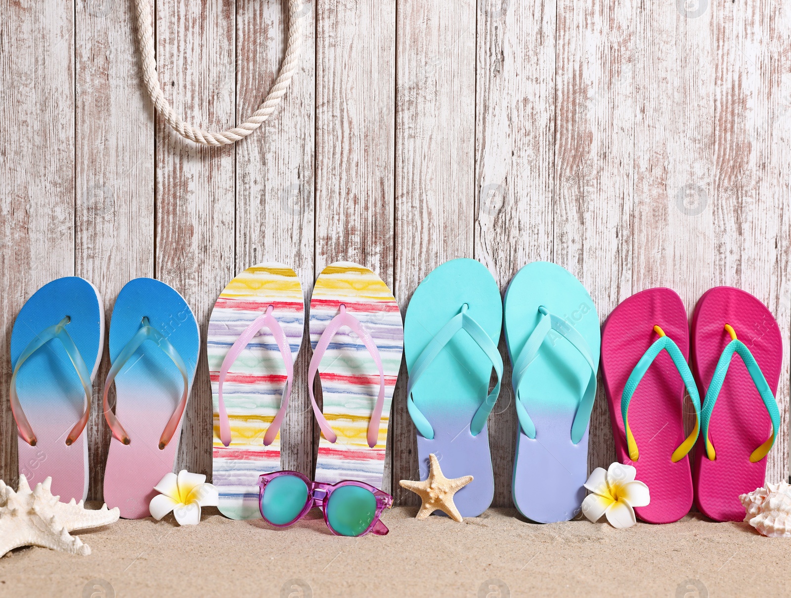 Photo of Different bright flip flops and sunglasses on sand near wooden wall, space for text. Summer beach accessories