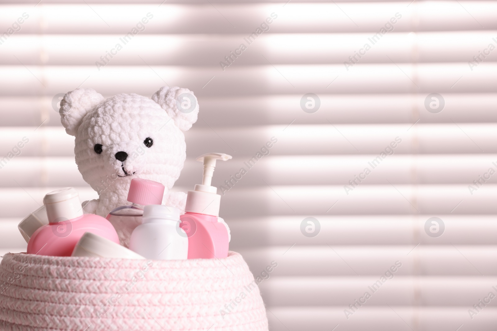 Photo of Wicker basket with baby cosmetic products and knitted toy bear near window, closeup. Space for text