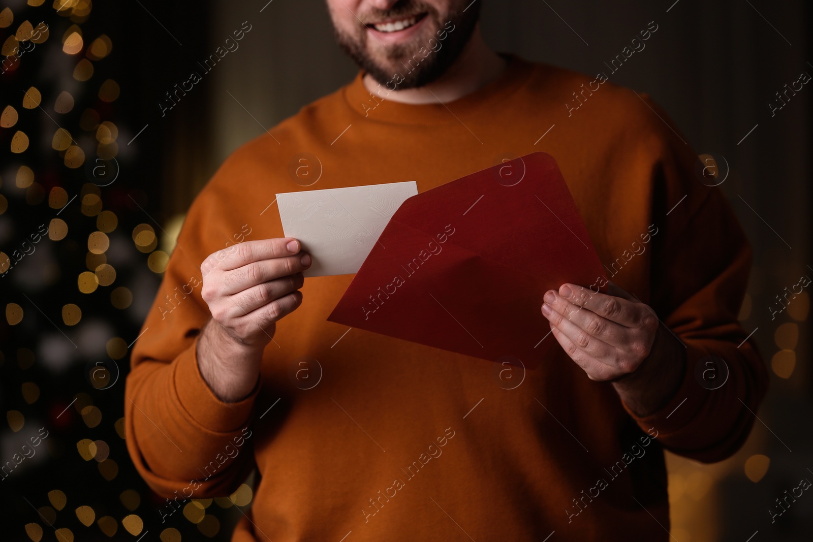 Photo of Man holding envelope and greeting card against blurred Christmas lights, closeup