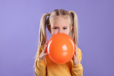 Cute little girl inflating orange balloon on violet background