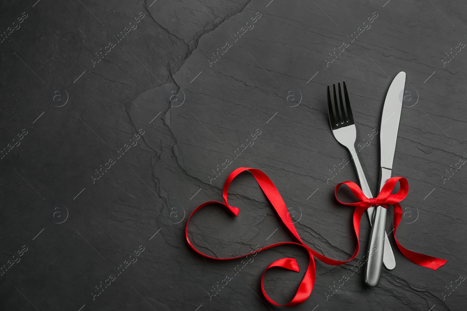 Photo of Cutlery set and red ribbon on black background, flat lay with space for text. Valentine's Day dinner