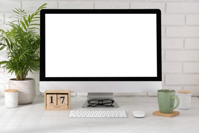 Photo of Office workplace with computer, glasses, cup, wooden block calendar and houseplant on light table near white brick wall