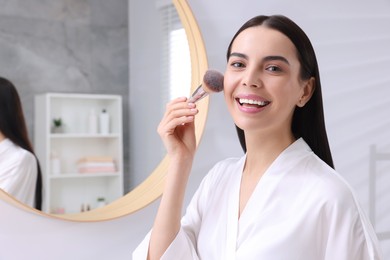 Beautiful young woman applying makeup with brush in bathroom. Space for text