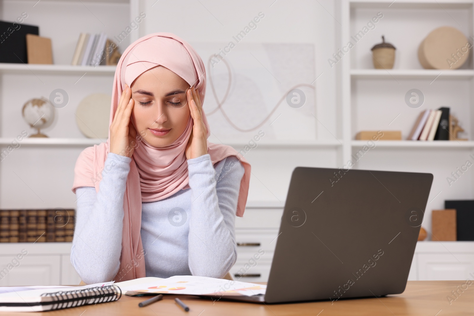 Photo of Tired Muslim woman in hijab working near laptop at wooden table in room