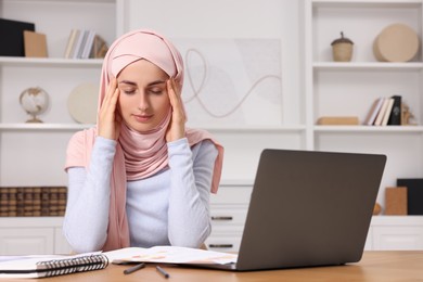 Tired Muslim woman in hijab working near laptop at wooden table in room