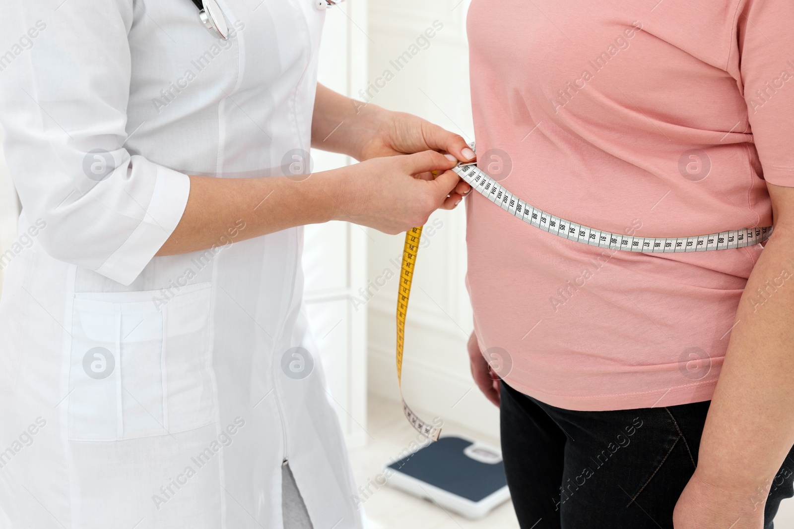 Photo of Nutritionist measuring overweight woman's waist with tape in clinic, closeup