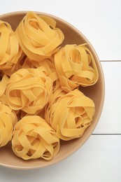 Raw tagliatelle pasta in bowl on white wooden table, top view