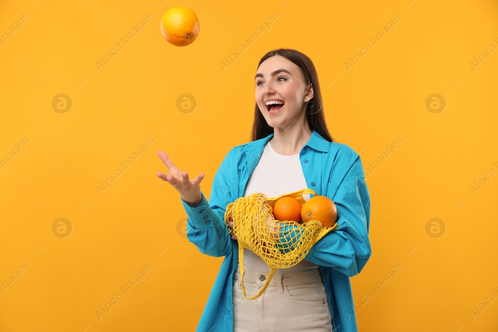 Photo of Woman with string bag of fresh oranges throwing fruit on orange background