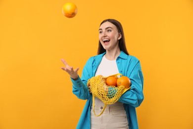 Woman with string bag of fresh oranges throwing fruit on orange background