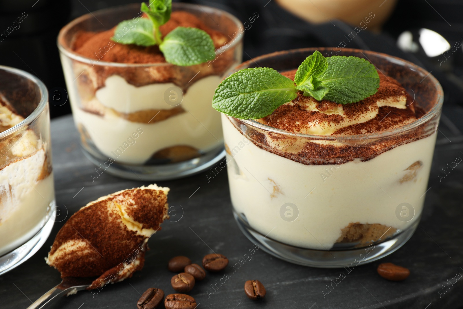 Photo of Delicious tiramisu in glasses, spoon, coffee beans and mint leaves on table, closeup