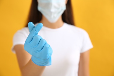Woman in protective face mask and medical gloves showing heart gesture on yellow background, closeup
