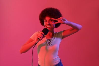 Curly young woman with microphone singing and showing victory sign on pink background. Color tone effect