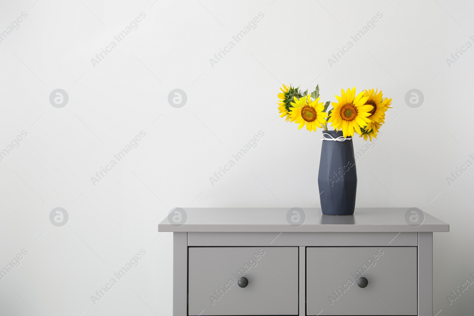 Photo of Bouquet of beautiful sunflowers in vase on commode near light wall. Space for text