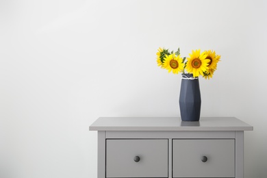 Photo of Bouquet of beautiful sunflowers in vase on commode near light wall. Space for text