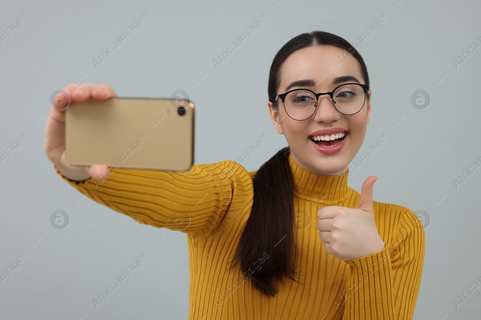 Photo of Smiling young woman taking selfie with smartphone and showing thumbs up on grey background