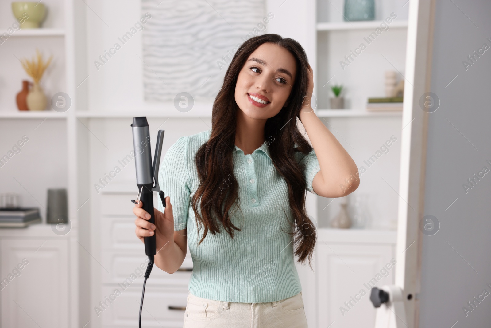 Photo of Smiling woman with beautiful hairstyle holding curling hair iron at home