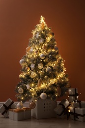 Beautiful decorated Christmas tree with glowing fairy lights and presents near orange wall indoors