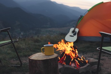 Photo of Yellow mug with hot drink on wooden stump near bonfire outdoors. Camping season