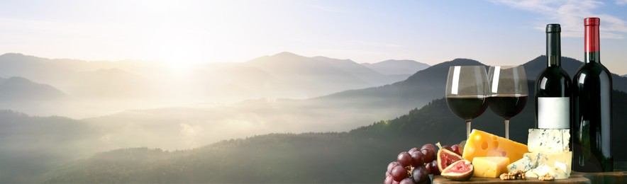 Tasty wine, grapes and cheeses on wooden table against beautiful mountain landscape, space for text. Banner design