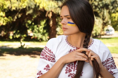 Sad young woman with drawing of Ukrainian flag on face in park, space for text