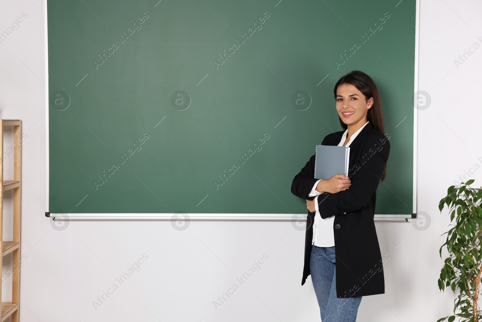 Photo of Happy young teacher with books at blackboard in classroom. Space for text