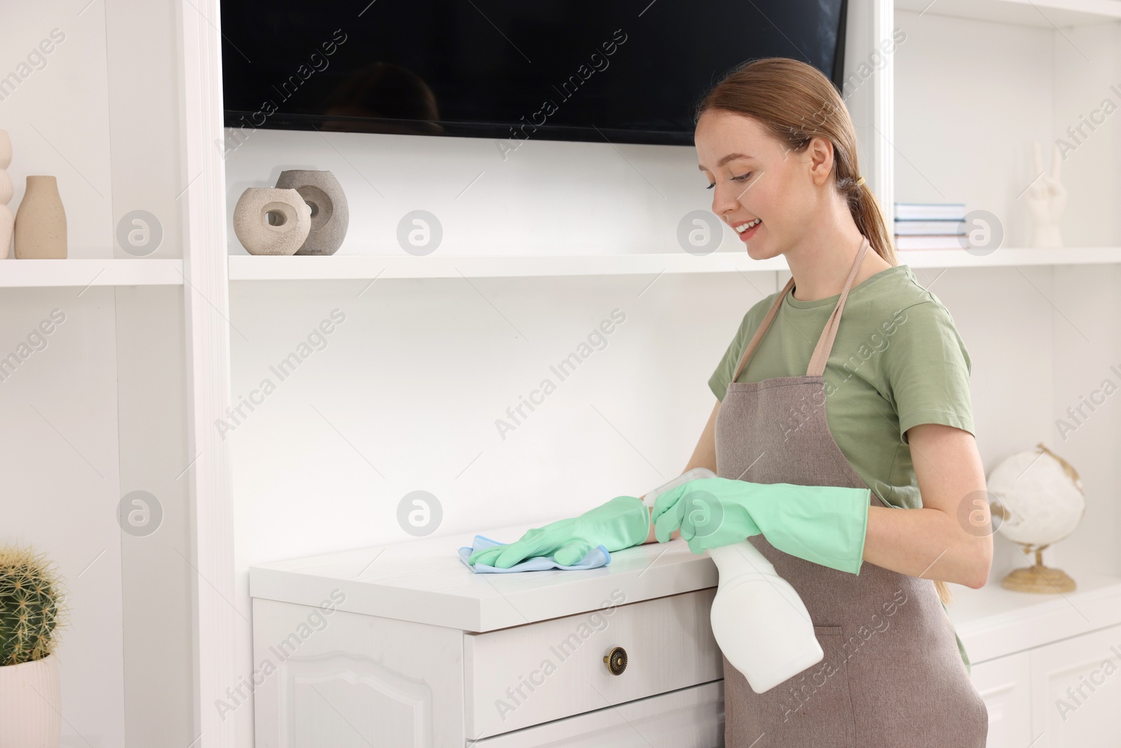 Photo of Woman with spray bottle and microfiber cloth cleaning white chest of drawers in room,