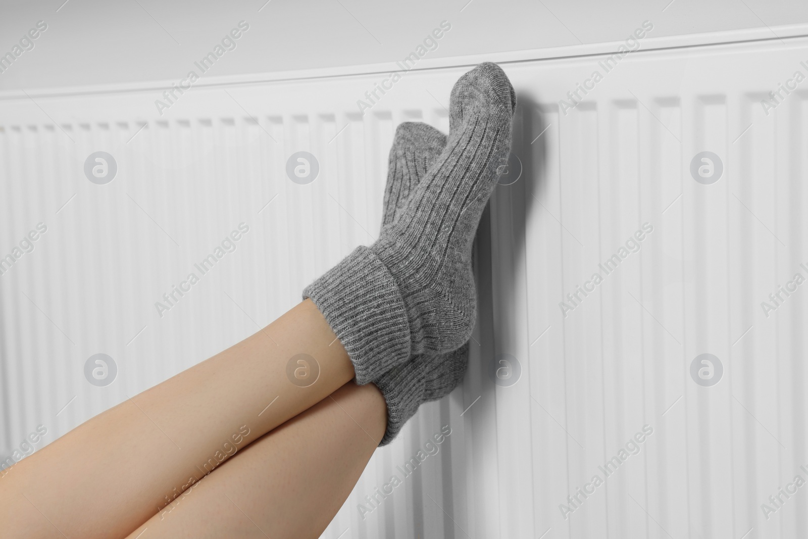 Photo of Woman warming legs on heating radiator near white wall, closeup