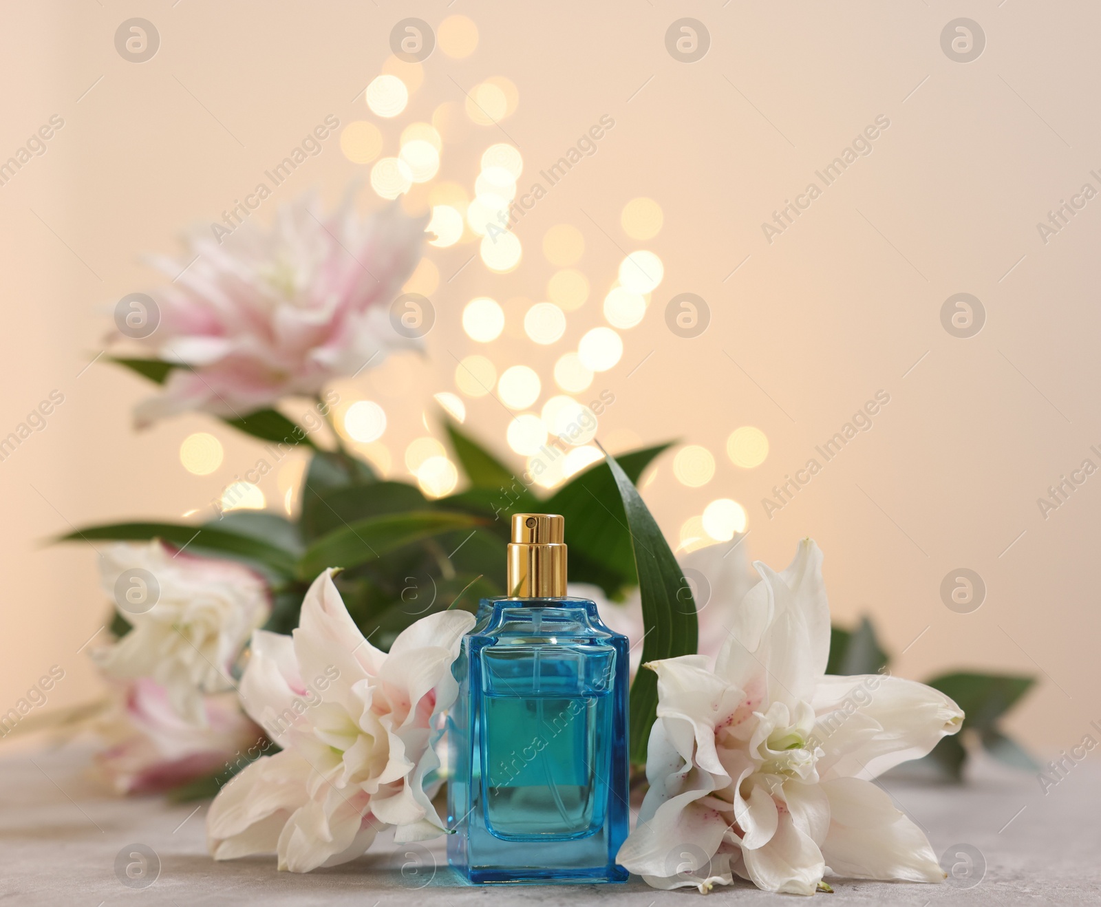 Photo of Bottle of perfume and beautiful lily flowers on table against beige background with blurred lights, closeup