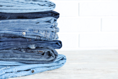 Photo of Stack of different jeans on white wooden table against brick wall. Space for text