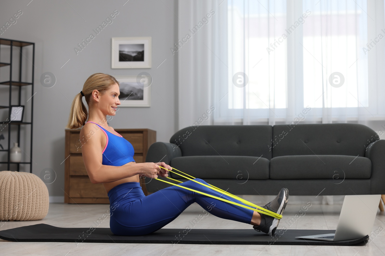 Photo of Fit woman doing exercise with fitness elastic band near laptop on mat at home