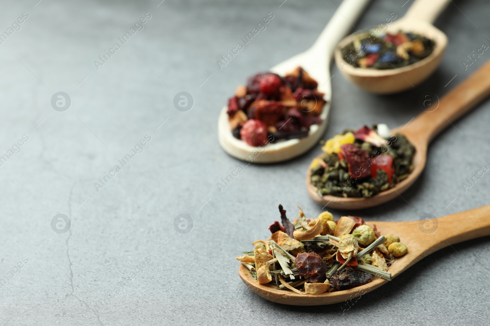 Photo of Different kinds of dry herbal tea in wooden spoons on light grey table, closeup. Space for text