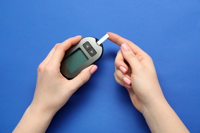 Photo of Diabetes. Woman checking blood sugar level with glucometer on blue background, top view