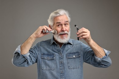 Senior man with mustache holding razor and brush on grey background