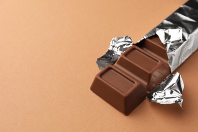 Photo of Delicious chocolate bar wrapped in foil on light brown background, closeup. Space for text