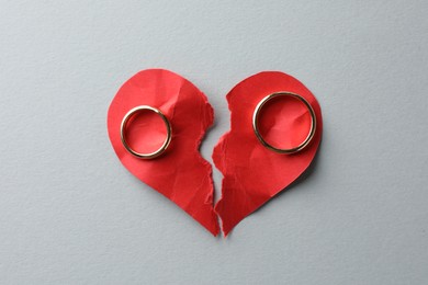 Photo of Halves of torn red paper heart and wedding rings on white background, top view. Broken heart