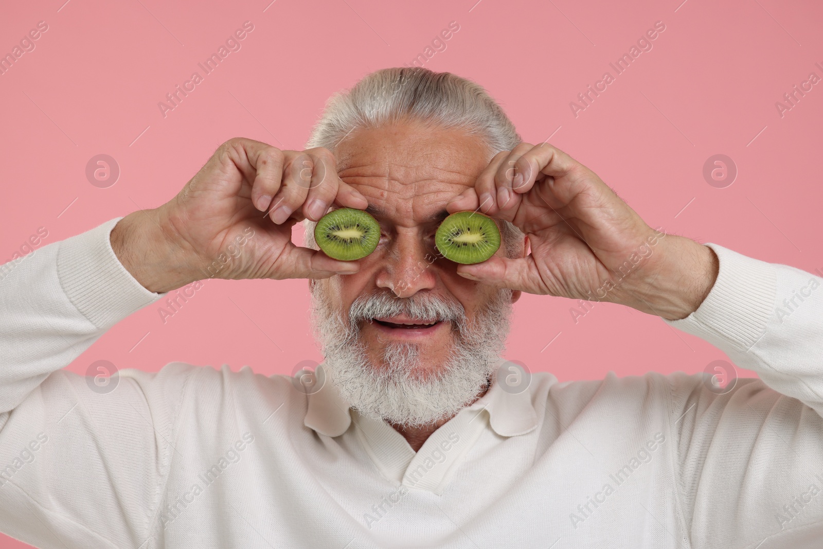 Photo of Senior man covering eyes with halves of kiwi on pink background