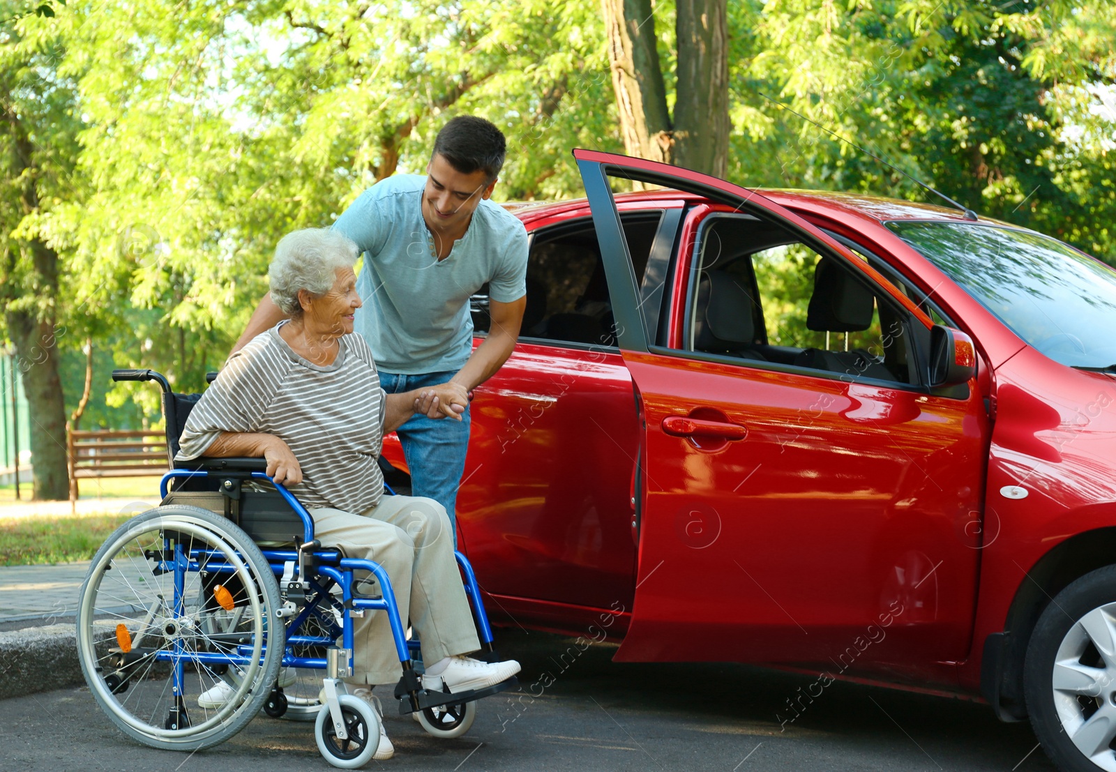 Photo of Young man helping disabled senior woman in wheelchair to get into car outdoors