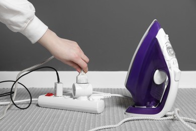 Photo of Woman putting plug from iron into power strip on grey carpet indoors, closeup