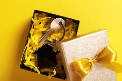 Key with trinket in shape of house and gift box on yellow background, flat lay. Housewarming party
