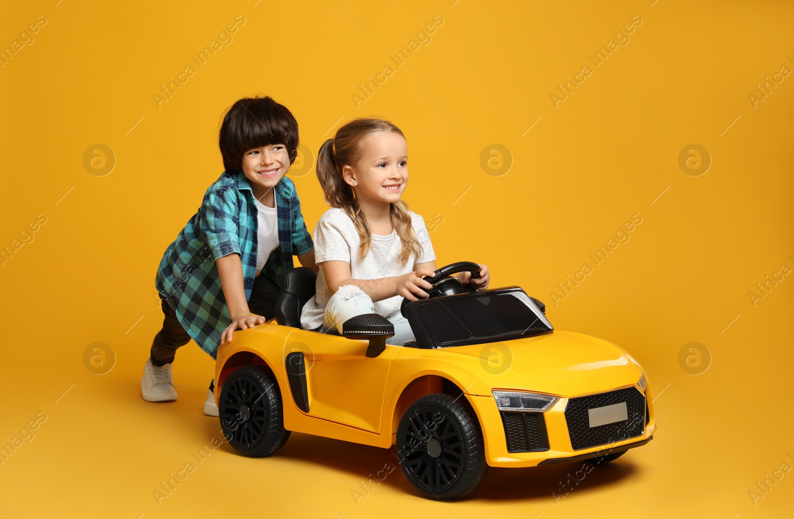 Photo of Cute boy pushing children's electric toy car with little girl on yellow background