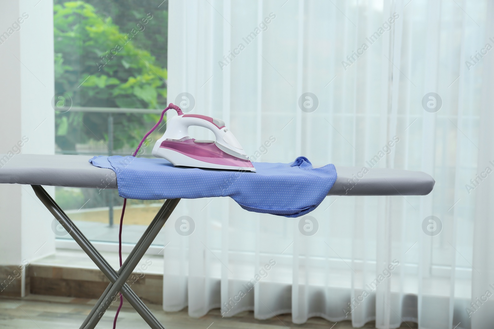 Photo of Modern iron and clean shirt on board near window indoors