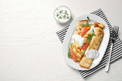 Delicious thin pancakes with salmon and sour cream on white wooden table, flat lay. Space for text