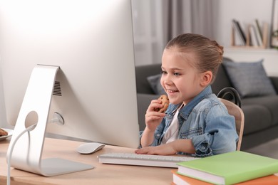 Photo of Little girl eating biscuit while using computer at table in room. Internet addiction